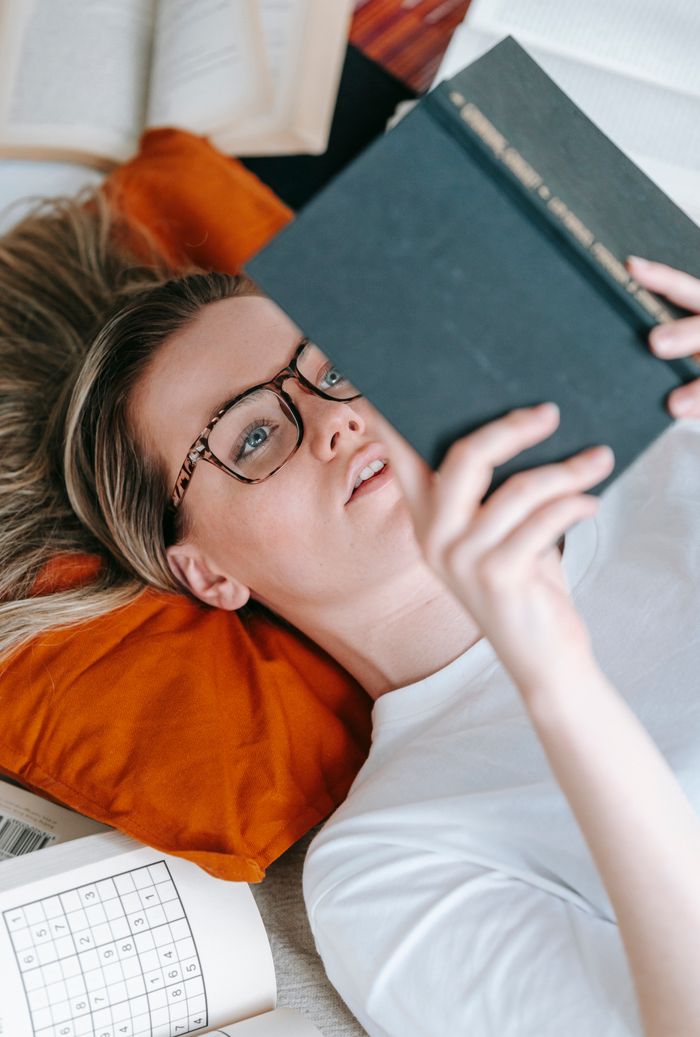 From above of serious female in eyeglasses lying on pillow amidst heap of opened books while reading novel in room